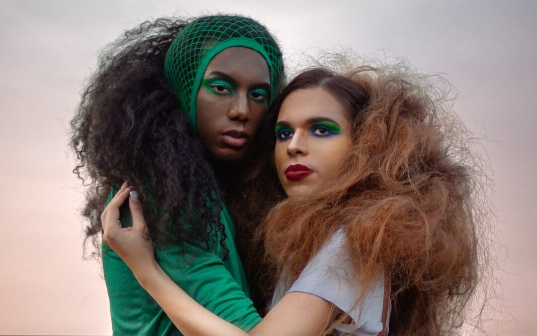 A Black femme wearing green with matching eye make-up holds a white-passing femme wearing grey with red lipstick and blue eye makeup.