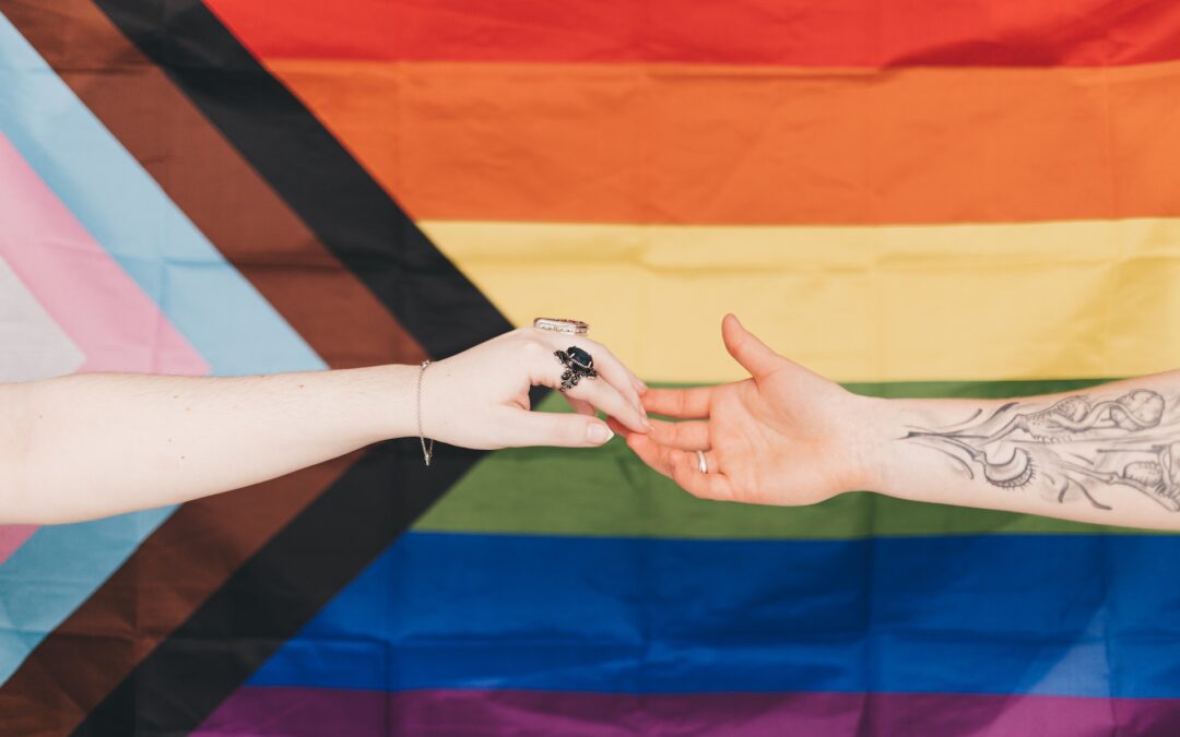 Queer pride rainbow flag with black, brown, and trans arrow stripes has two white hands touching each other in the foreground