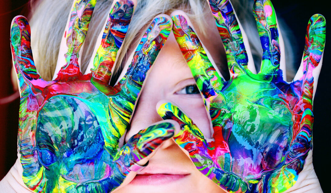 A child against a black background holds up palms smeared with rainbow paint in front of their face.