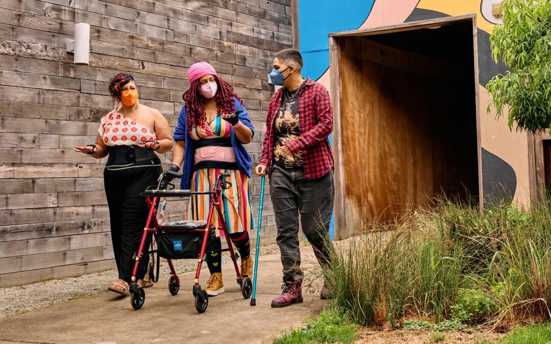 Three masked disabled people of color chat while strolling through an outdoor courtyard. On the left, an Indigenous woman wears glasses, wrist support braces, and waist trainers paired with a off-shoulder top and wide leg pants. In the middle, a Black non-binary woman walks with a rollator and wears waist trainers with ice pack inserts over their rainbow striped dress. On the right, a Latinx non-binary person walks while using a teal functional grip cane.