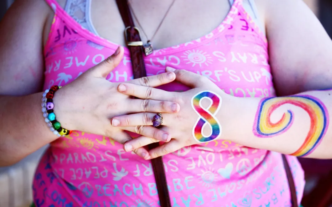 A feminine person wearing a pink shirt holds their hands on their chest. They have a rainbow infinity symbol painted on their hand.